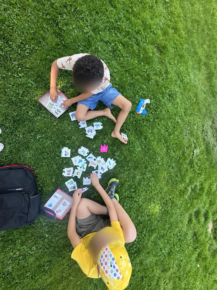 Two children lying on the grass outdoors, playing a matching card game together. Faces are blurred.